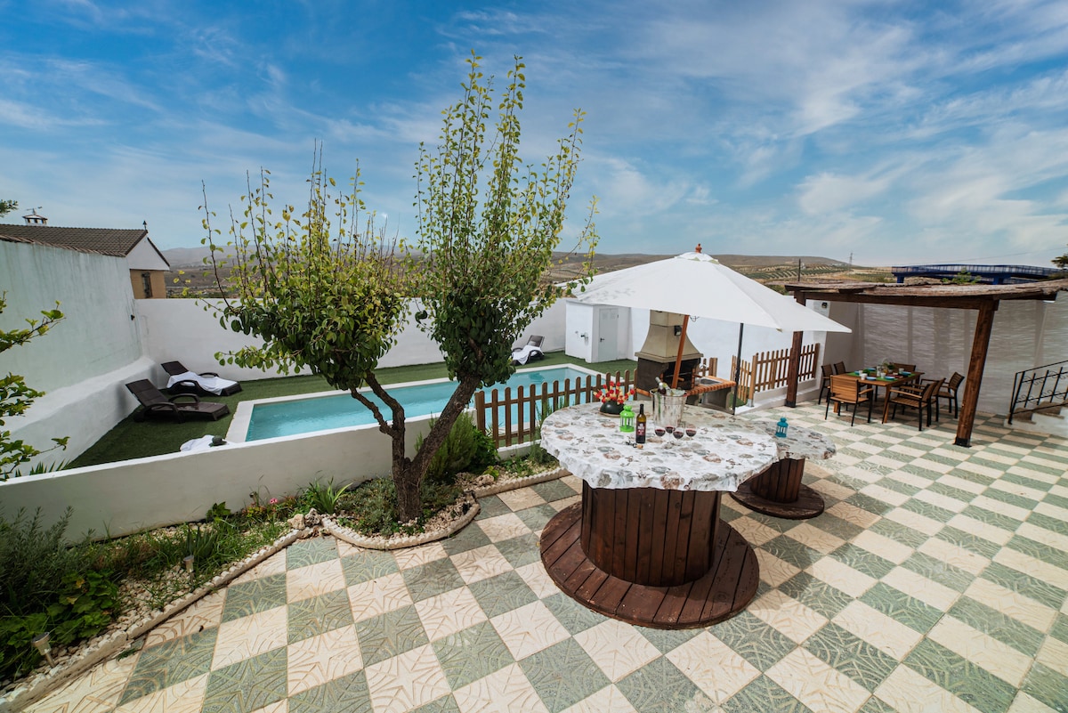 An outdoor area features two private pools surrounded by a secure fence and a shaded dining space. A barbecue station is positioned near a tree, while lounge chairs are arranged around the pools. The ground is decorated with patterned tiles, enhancing the outdoor setting.