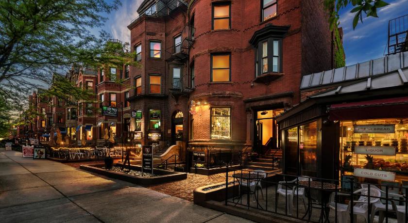 A charming street in Back Bay features elegant brownstone buildings with large windows and detailed brickwork. Restaurant patios with tables and chairs line the sidewalk, inviting guests to enjoy the neighborhood's vibrant atmosphere. A gentle glow from buildings indicates evening activity.