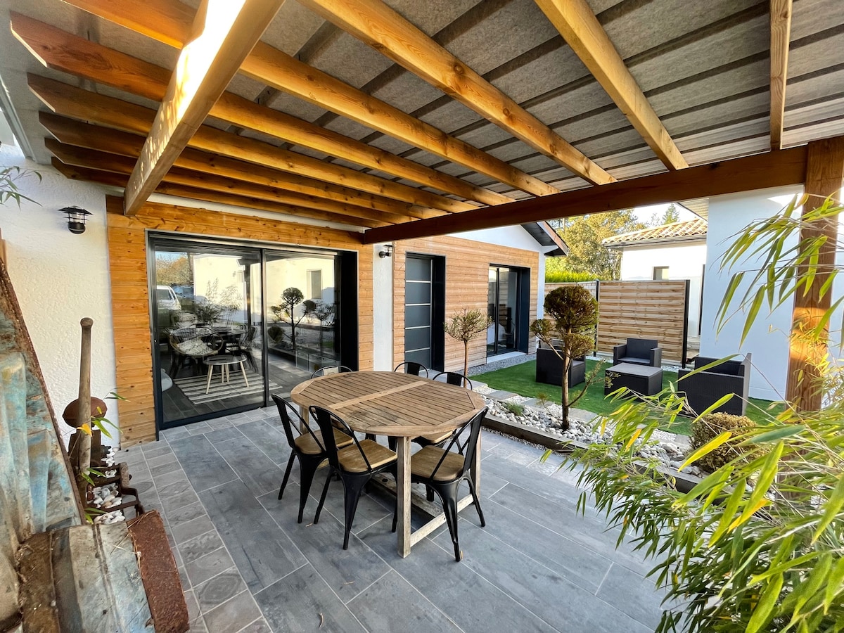 A covered terrace with wooden beams features a round dining table surrounded by six chairs. The space is adjacent to a small private garden, with landscaped areas visible. Glass sliding doors provide access to the interior living area, enhancing the connection between indoor and outdoor spaces.