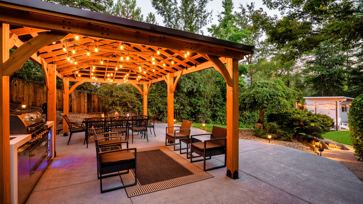 An outdoor living space is showcased under a wooden pavilion adorned with string lights. A barbecue grill is positioned nearby, complemented by multiple seating areas featuring comfortable chairs and a table. Lush greenery surrounds the space, enhancing the serene atmosphere.