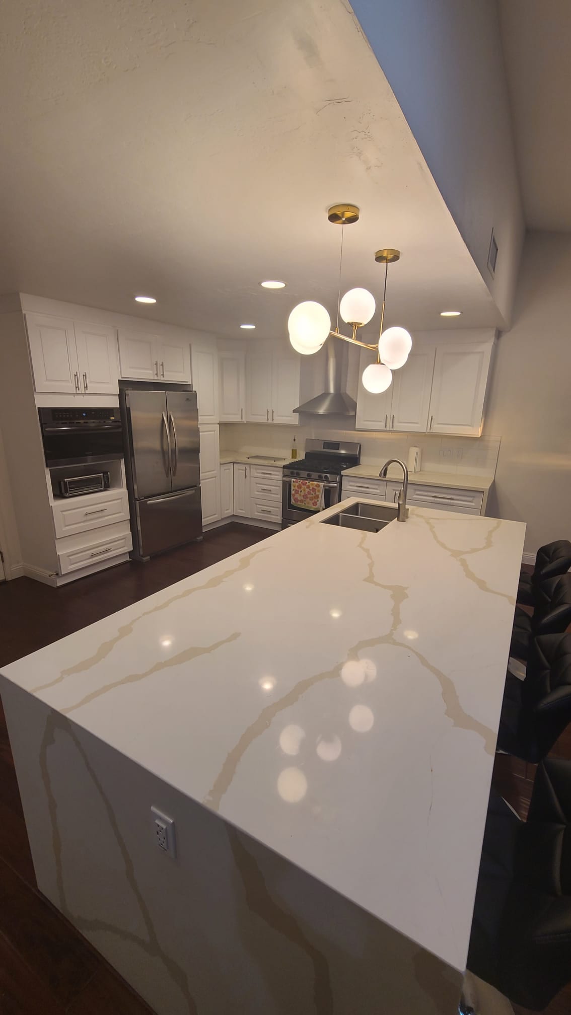 A modern kitchen features a large island with a white countertop marked by subtle golden veining. The cabinetry is white, complemented by stainless steel appliances. Pendant lighting above the island adds a contemporary touch, while warm lighting creates an inviting atmosphere.