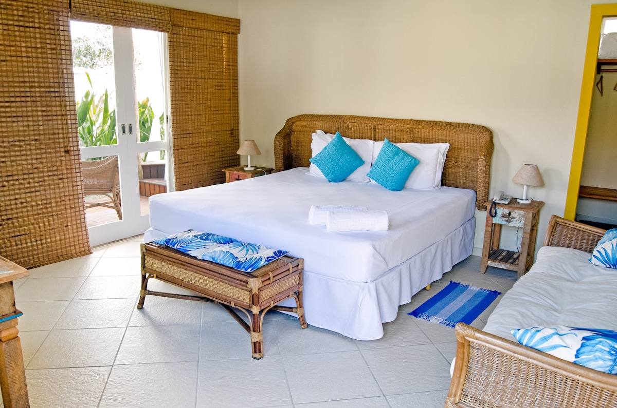 A spacious sleeping area features a large bed adorned with decorative cushions. Woven furniture adds texture, while natural light filters through sliding doors that open to a private patio. A small bedside table and lamp are positioned beside the bed.