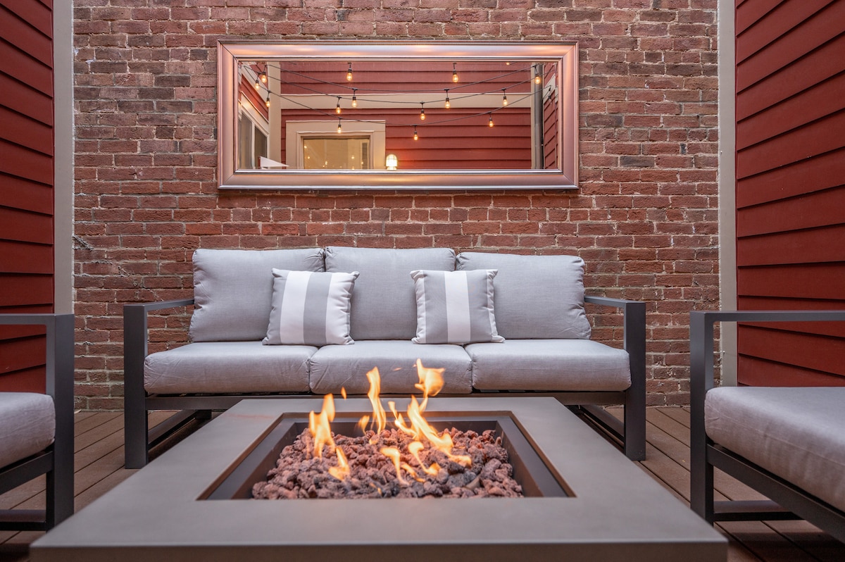 An outdoor seating area features a modern fire table surrounded by a comfortable gray sofa adorned with soft cushions. A large mirror reflects the inviting atmosphere, while the warm glow of the fire adds a cozy touch. Exposed brick walls and string lights complete the setting.