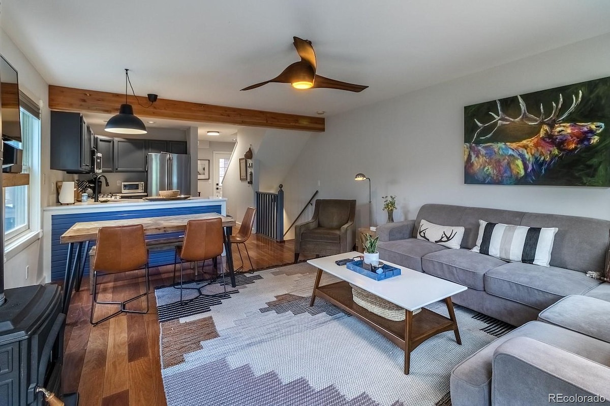 The spacious living area showcases contemporary decor with a large sectional sofa and a coffee table at its center. A dining area with metal chairs is positioned nearby, while a modern ceiling fan and artwork are featured on the walls. Bright light enters through large windows.