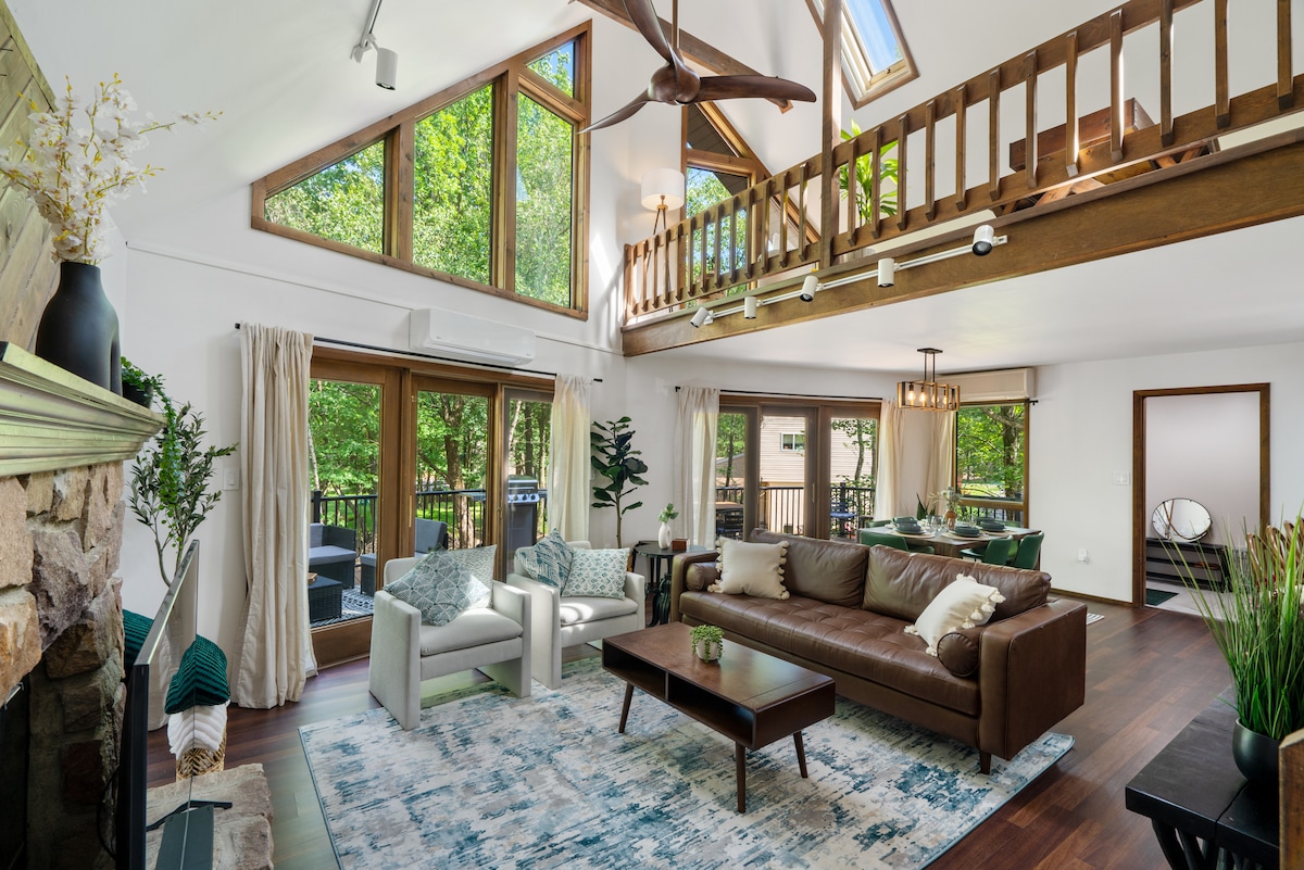 The spacious living area features large floor-to-ceiling windows allowing natural light to fill the room. A leather sofa and two white chairs are arranged around a dark coffee table on a patterned rug. A lofted space is visible above, with wooden railings overlooking the area.
