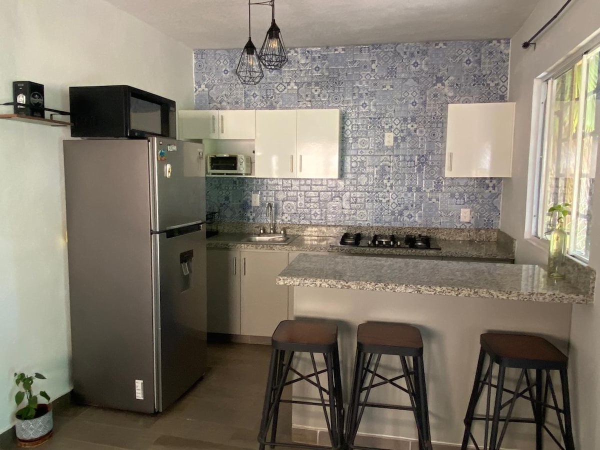 A modern kitchen is showcased with blue patterned tile backsplash. A stainless steel refrigerator and a microwave are present alongside white cabinetry. A granite countertop provides ample workspace, and three bar stools are positioned at the counter.