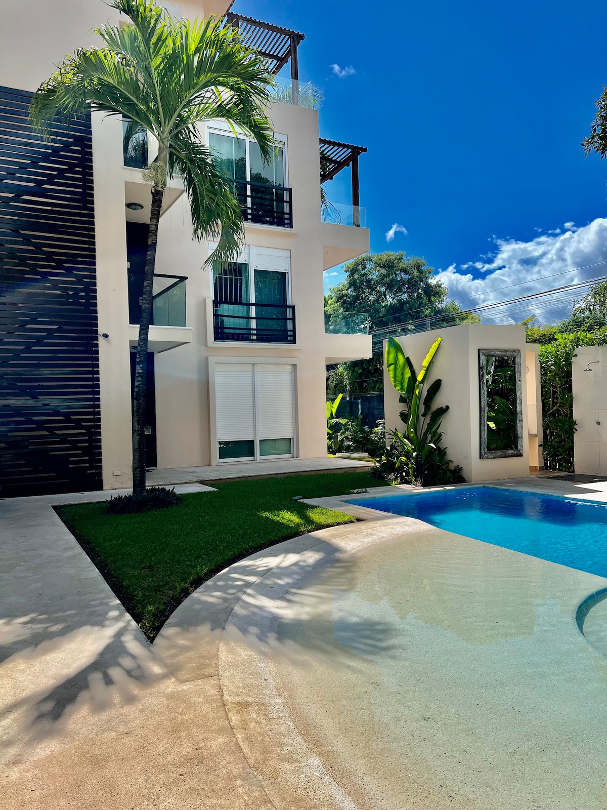 The image showcases a modern condominium building surrounded by lush greenery and a tranquil pool. The pool area is complemented by manicured grass and tropical plants, with sunlight reflecting off the water's surface, adding to the inviting ambiance of the outdoor space.
