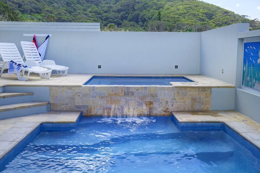 A contemporary pool area features a spacious swimming pool and a connected spa. Light blue tiles complement the natural surroundings, while a mural of marine life decorates one wall. Multiple lounge chairs are arranged around the pool for relaxation.