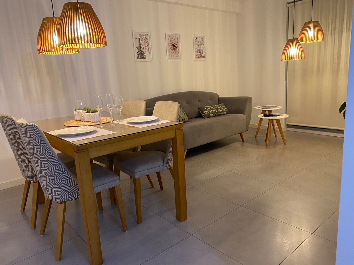 An elegantly arranged dining area features a wooden table set for four beside a comfortable gray sofa. Three pendant lights hang above, casting a warm glow. Light-colored chairs with textured fabric complement the sleek decor, while framed art pieces adorn the wall.