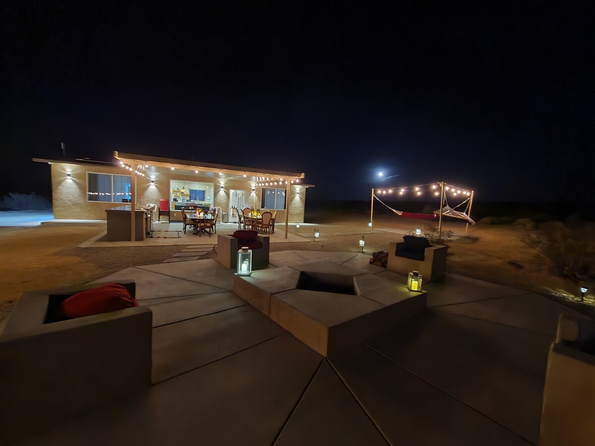 An outdoor area illuminated by Edison lights features a dining table and seating, with a custom fire pit surrounded by cozy cushions. A hammock area is visible in the background, creating a welcoming space for evening relaxation.