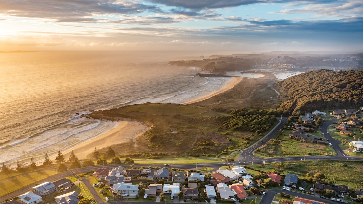 Aerial view showcases a serene coastline, highlighting gently rolling waves meeting sandy beaches. Lush greenery outlines the shore, with a blend of residential areas visible in the foreground. The scene is complemented by soft golden light, reflecting off the water and illuminating the landscape.