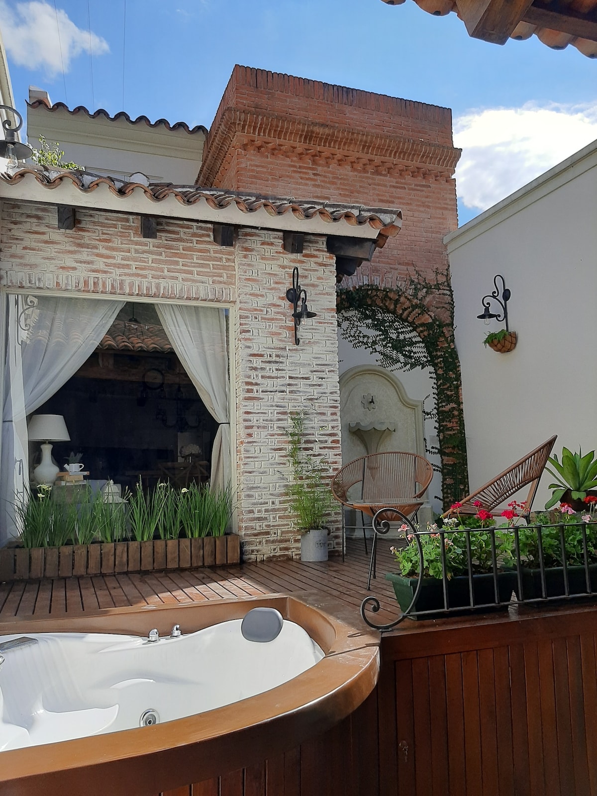 An outdoor deck features a wooden hot tub surrounded by greenery and potted plants. Soft curtains frame an adjoining living area, while rustic brick walls add character. Natural light filters in, enhancing the serene atmosphere of the space.