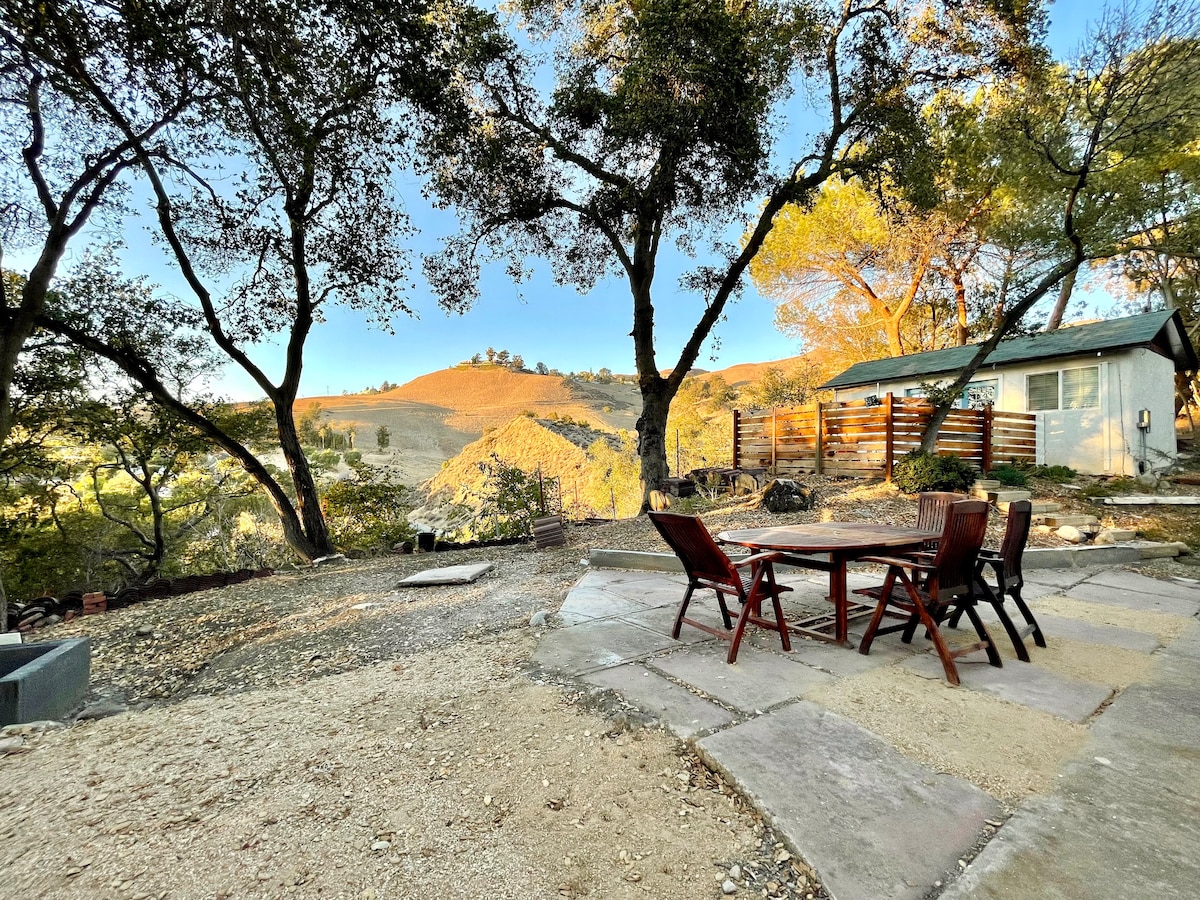 An outdoor seating area features a wooden table surrounded by four chairs, set on a stone patio. Sunlight filters through large trees, illuminating the natural landscape and distant hills. A rustic structure is visible in the background, enhancing the serene ambiance.