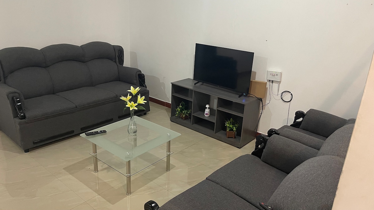 A living area is equipped with a gray sectional sofa and matching armchairs, providing comfortable seating. A glass coffee table sits in the center, accentuated by a vase with yellow flowers. A flat-screen TV is mounted on the wall with a media console below.