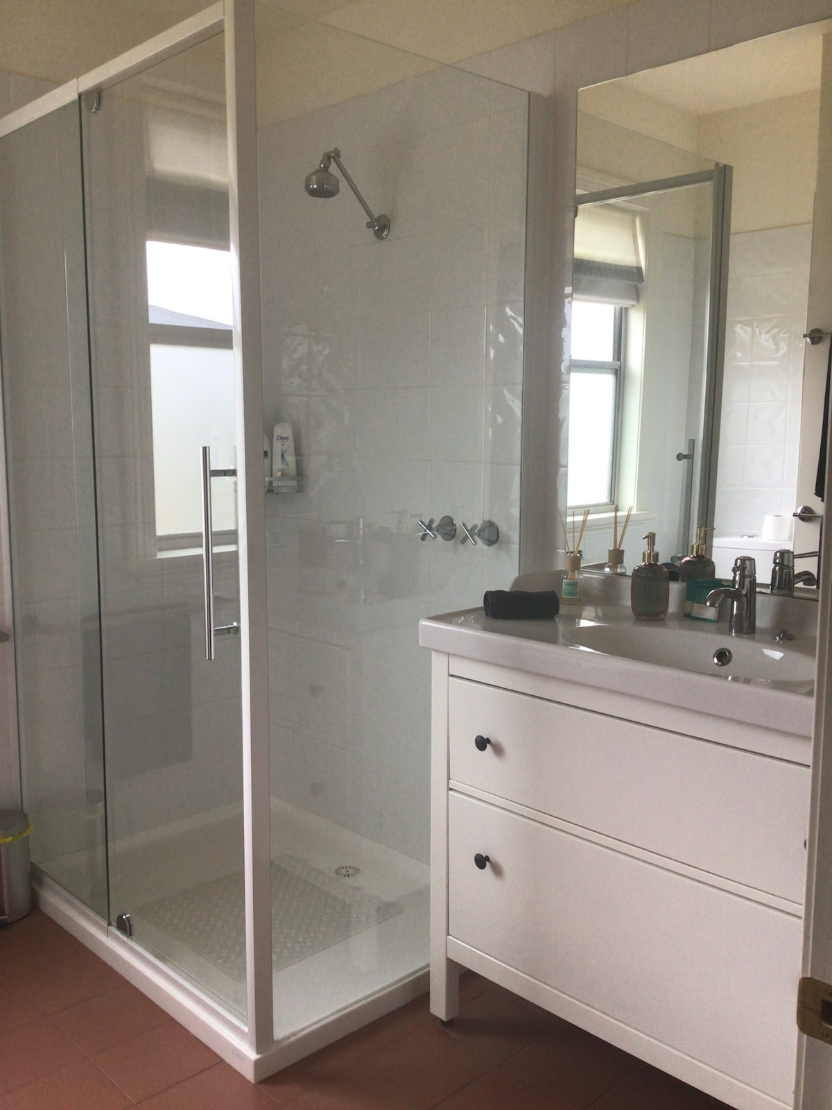 A modern bathroom features a glass shower enclosure with a sleek showerhead. A vanity with a smooth white surface offers several bathroom accessories. Natural light enters through a nearby window, which is framed by a large mirror, enhancing the sense of space.