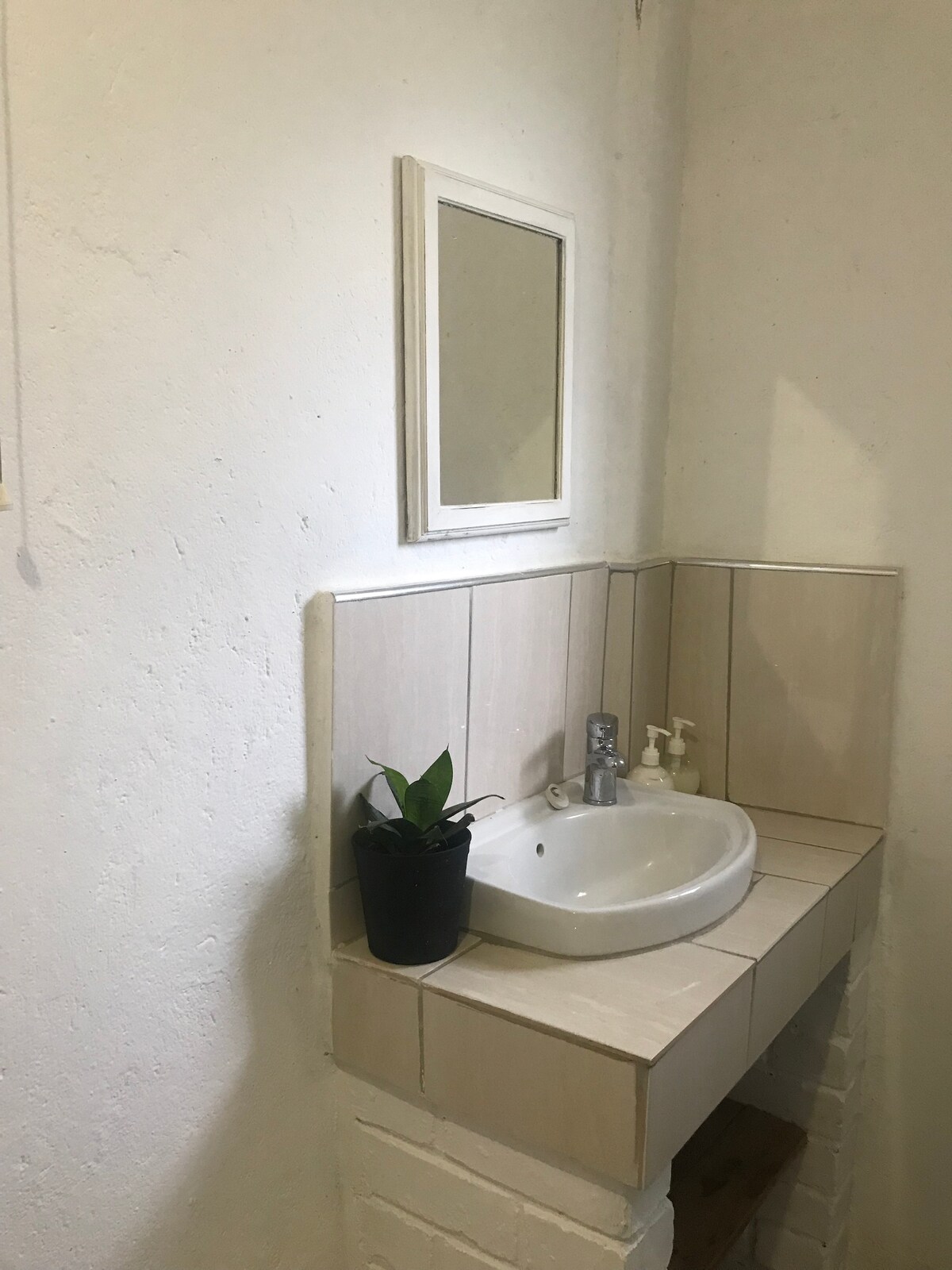A small bathroom area features a white basin set into beige tiled counters. A simple mirror hangs above the basin, and a small potted plant sits beside it. The walls are painted a light color, enhancing the bright and clean look of the space.