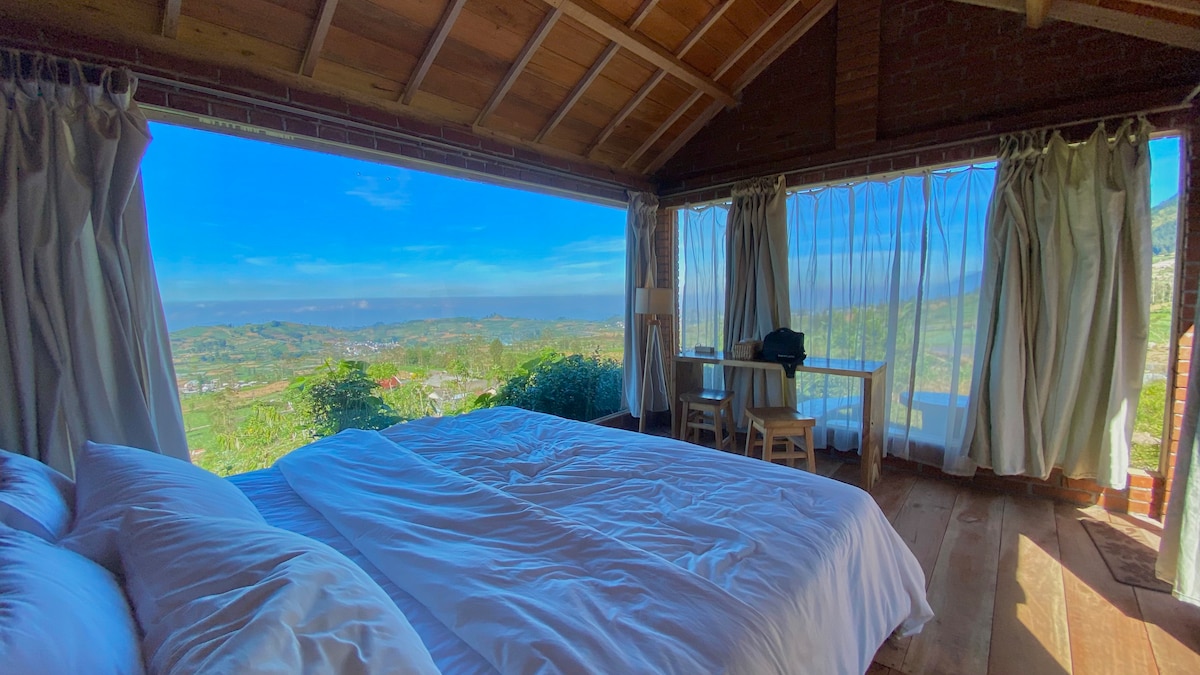 A spacious bedroom features a large bed with white linens, complemented by generous windows that frame panoramic views of the hills and valley. Natural light floods the room, enhancing the wooden floor and simple furniture, including a small table and chairs positioned near the glass doors.