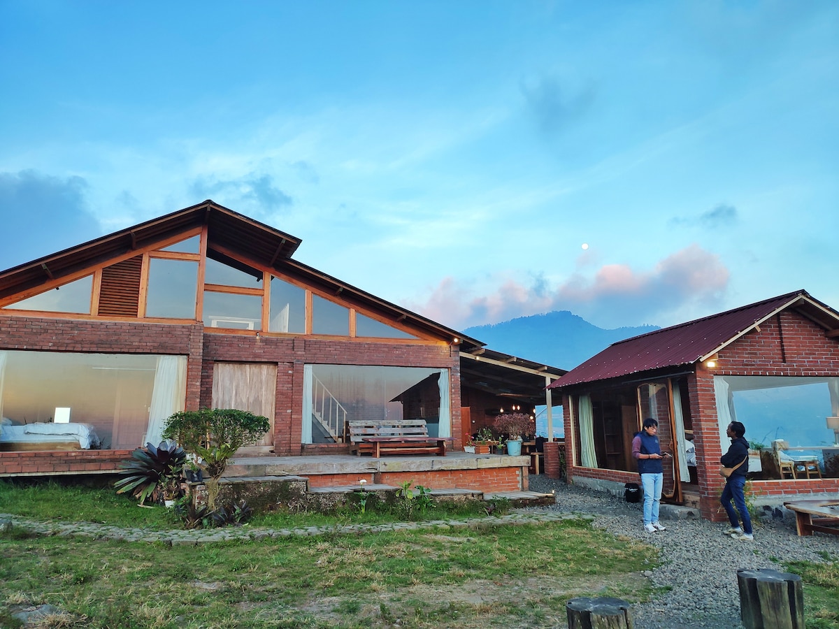 The exterior of a spacious brick villa is highlighted against a dusky sky, featuring expansive glass windows that reflect the surrounding landscape. A wooden deck extends outside, with seating arrangements and lush greenery. Two individuals are seen enjoying the fresh air beside the structure.