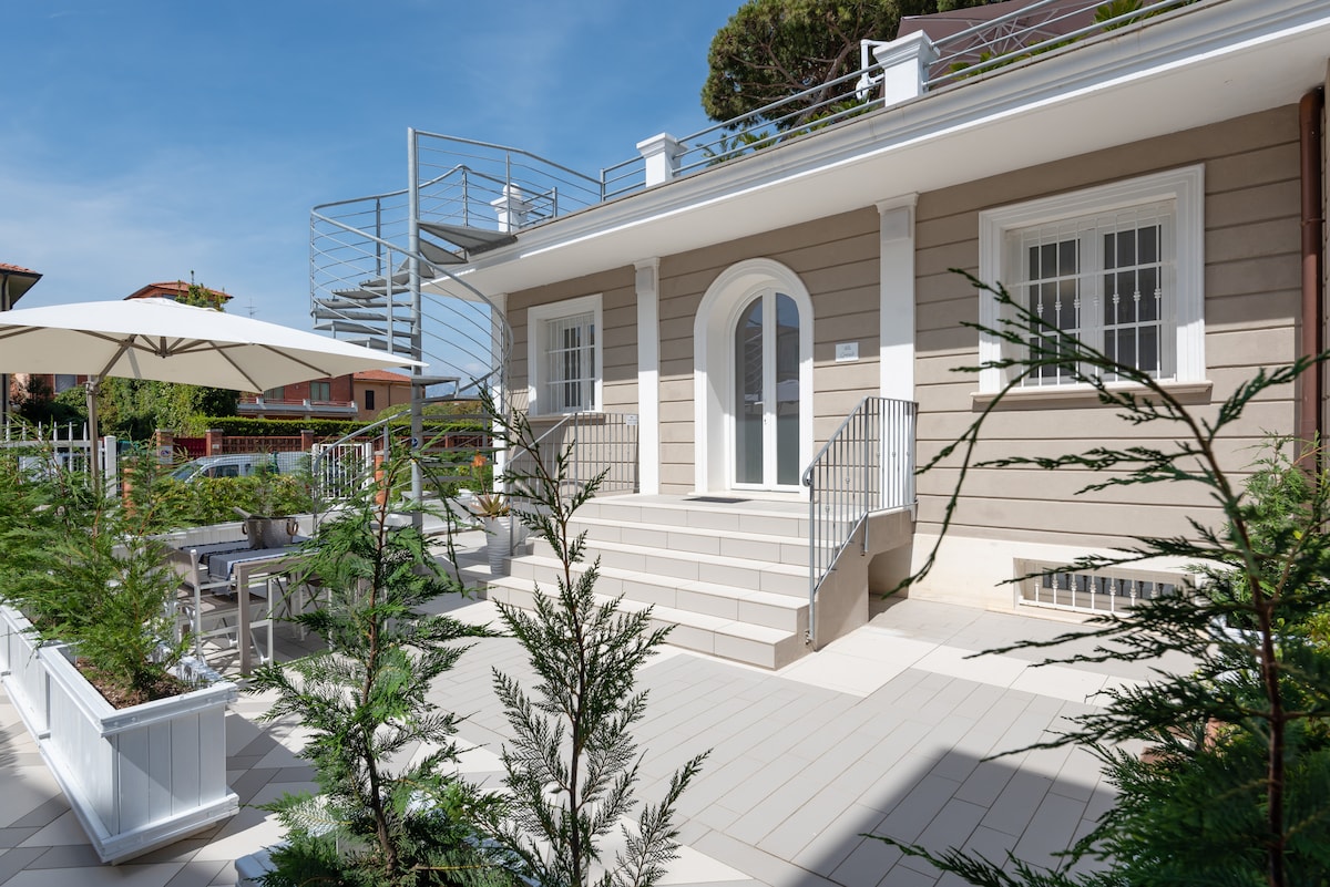 The villa features a welcoming entrance with a broad staircase leading to an elevated porch. Light-colored walls and clean lines are complemented by neatly arranged potted plants. An outdoor dining area with an umbrella provides a comfortable setting for relaxation.
