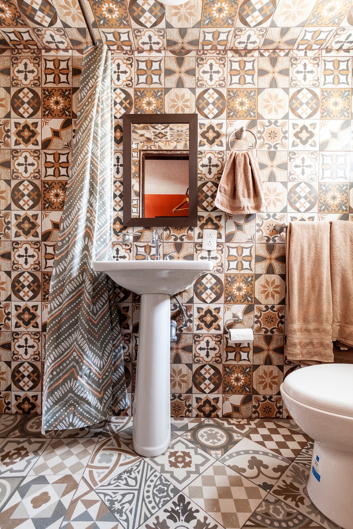 A bathroom features decorative patterned tiles on the walls and flooring. A white pedestal sink is positioned under a square mirror. A towel hangs on a bar beside the sink, and a toilet is visible, completing the functional layout of the space.