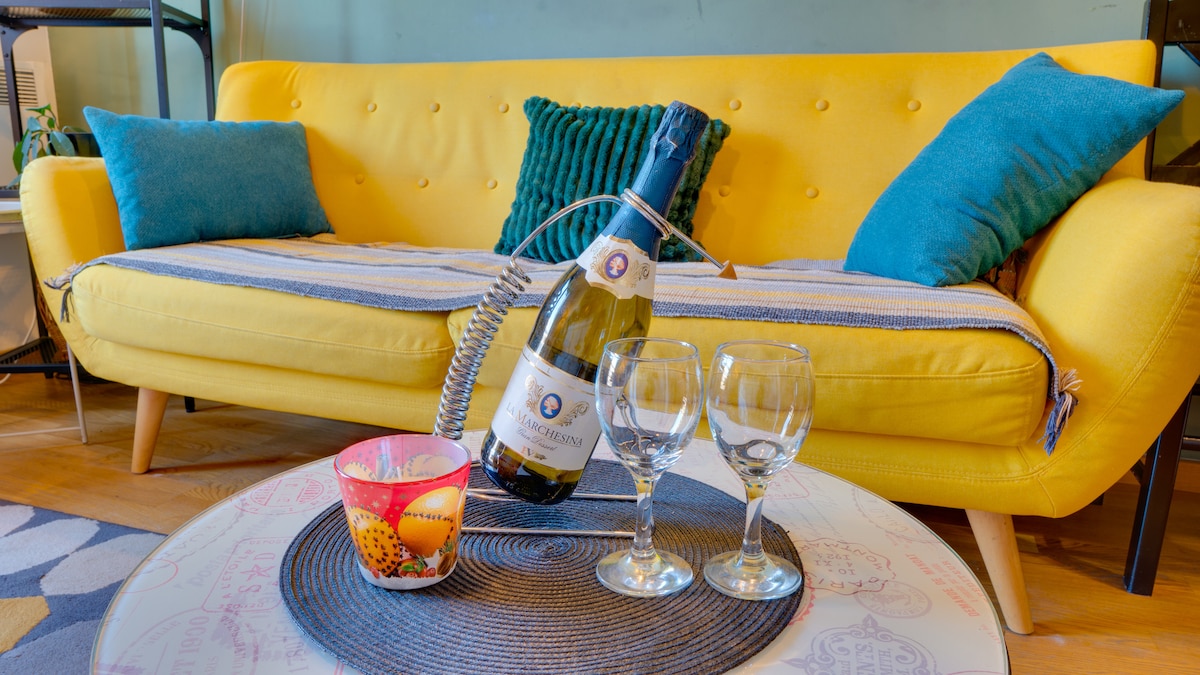 A vibrant yellow sofa is complemented by teal cushions, creating a bright seating area. A bottle of champagne sits on a round table beside two glasses. A decorative candle adds a touch of warmth to the setting, enhancing a welcoming atmosphere for relaxation.