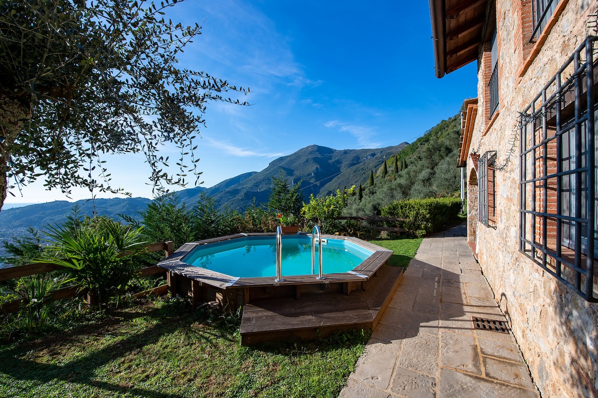 A wooden deck surrounds a circular swimming pool, offering a serene view of the surrounding mountains and greenery. The rustic cottage is visible beside the pool, showcasing local stone and warm-toned brick. Lush plants frame the tranquil outdoor space.