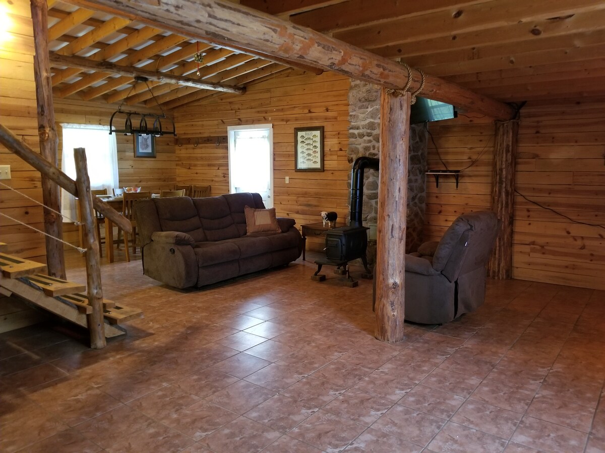The lodge interior is characterized by wooden walls and beams, creating a cozy ambiance. A spacious seating area features two comfortable couches arranged around a small wood stove. A dining table is visible in the background, complemented by natural light filtering through the windows.