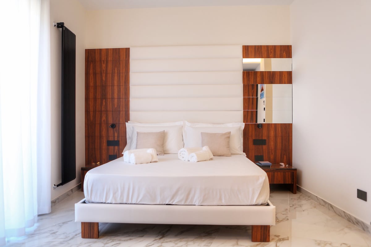 A spacious double bed is centered in the room, complemented by wooden headboards and bedside tables. Light-colored linens and multiple towels are arranged neatly on the bed. A large mirror adorns the wall beside the bed, reflecting natural light from nearby windows.
