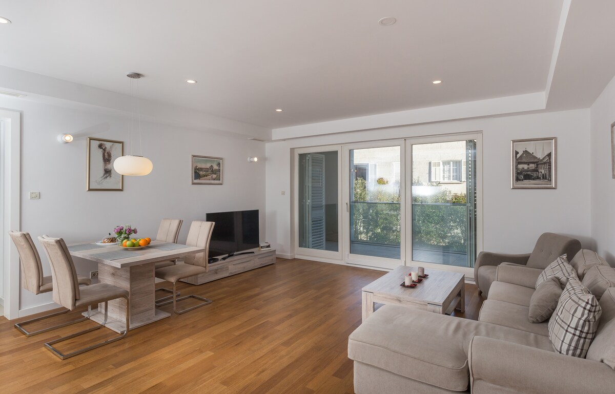 A spacious living area features a comfortable sofa and a dining table for six. Large glass doors open to a balcony with greenery visible outside. A television is positioned against a wall, and framed artwork decorates the walls, enhancing the modern aesthetic.