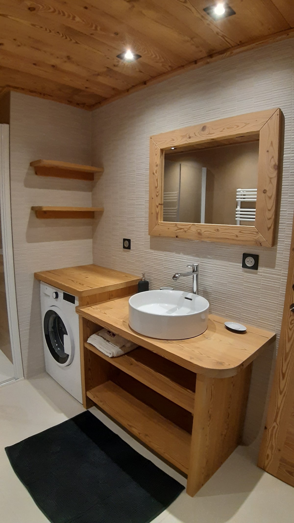 A modern bathroom features wooden accents with a round sink placed atop a wooden countertop. Shelves are installed above the counter for additional storage, while a washing machine is situated beside the countertop. Soft lighting highlights the wood ceiling and textured wall.