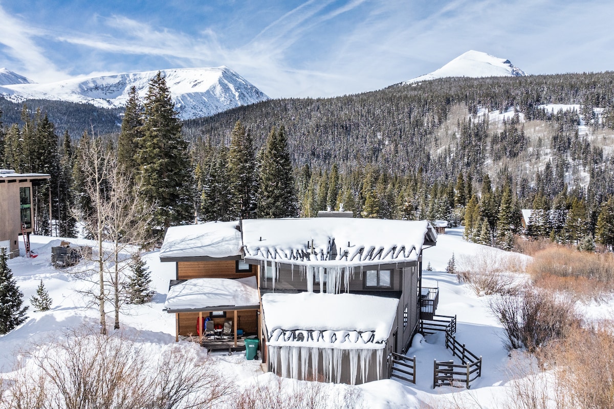 A snow-covered exterior of a multi-level home is displayed, featuring a large roof with icicles. The house is surrounded by a snowy landscape and pine trees, with views of distant mountains rising in the background.