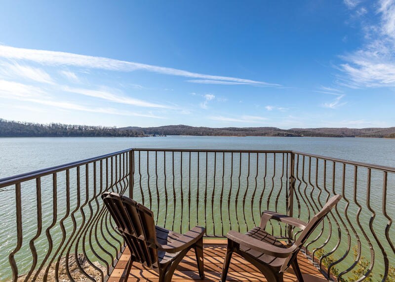A wooden deck overlooks Table Rock Lake, featuring two brown Adirondack chairs positioned for scenic views. The tranquil lake reflects the clear blue sky, while the surrounding hills are visible in the distance, creating a peaceful lakeside setting.