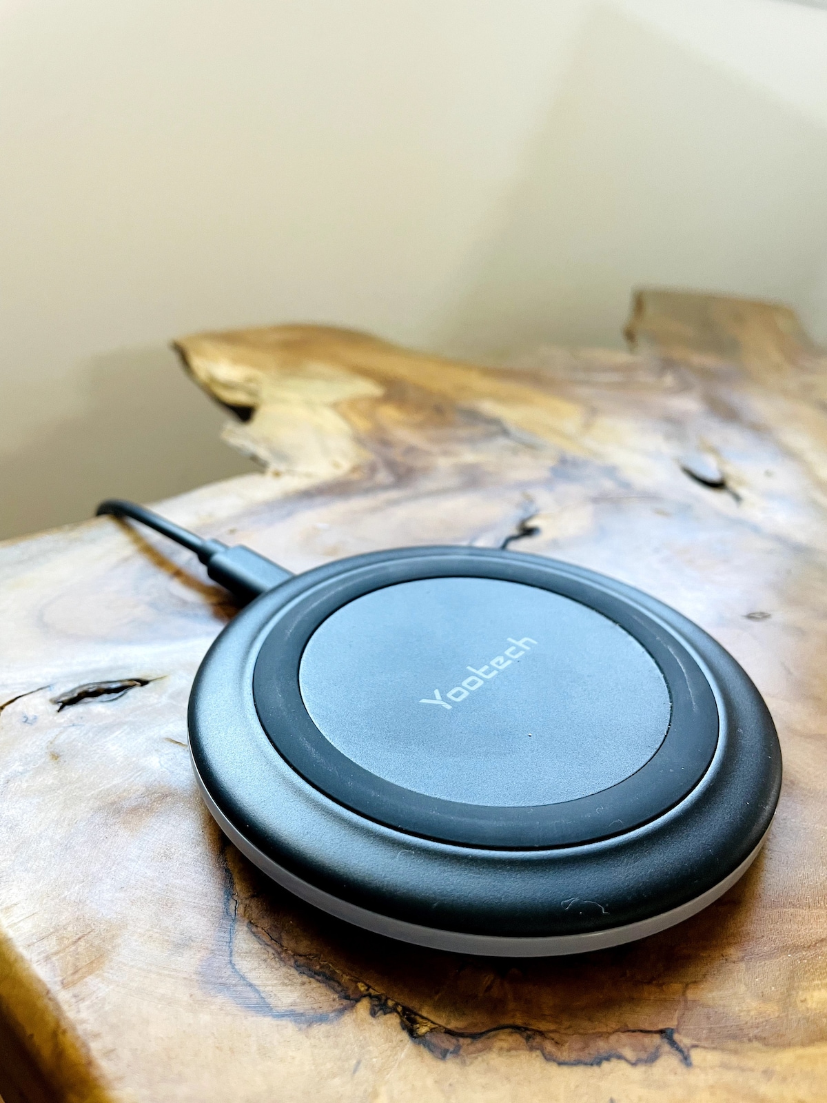 A wireless phone charger is placed on a textured wooden surface. The charger features a smooth circular design with a black finish, displaying the brand name in a subtle manner.