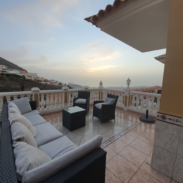 A terrace area is presented with a comfortable seating arrangement featuring a dark wicker sofa and two matching chairs. A small table is positioned at the center. The background captures a scenic view of the landscape and sky during sunset.
