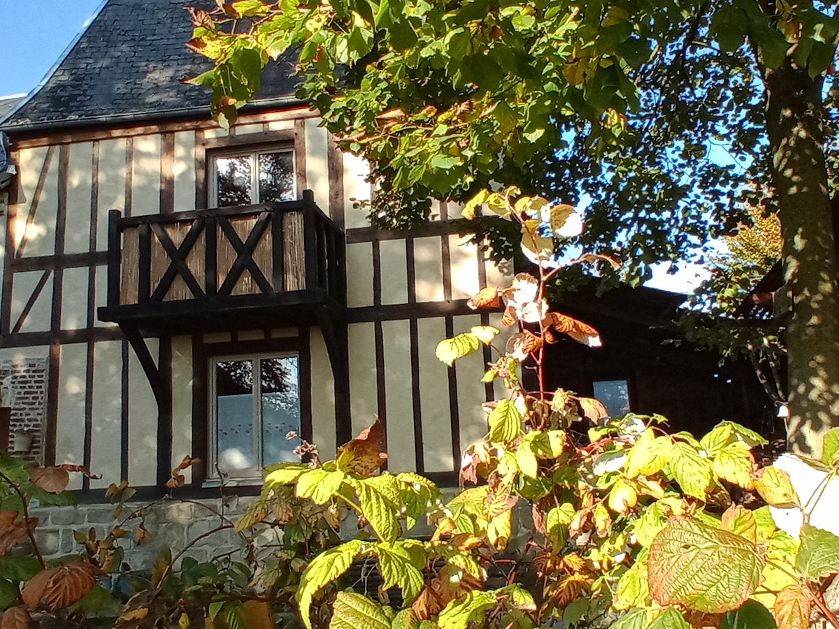 The gîte features a charming half-timbered design with a balcony overlooking vibrant greenery. Sunlight filters through the leaves of surrounding trees, highlighting the exterior details and creating a warm atmosphere in the garden.
