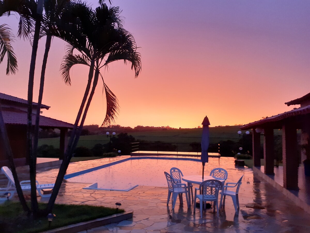 The outdoor area features a paved patio surrounding a rectangular pool, reflecting the colors of a sunset sky. Several palm trees provide shade, and a table with four chairs is set nearby, creating an inviting space for relaxation.
