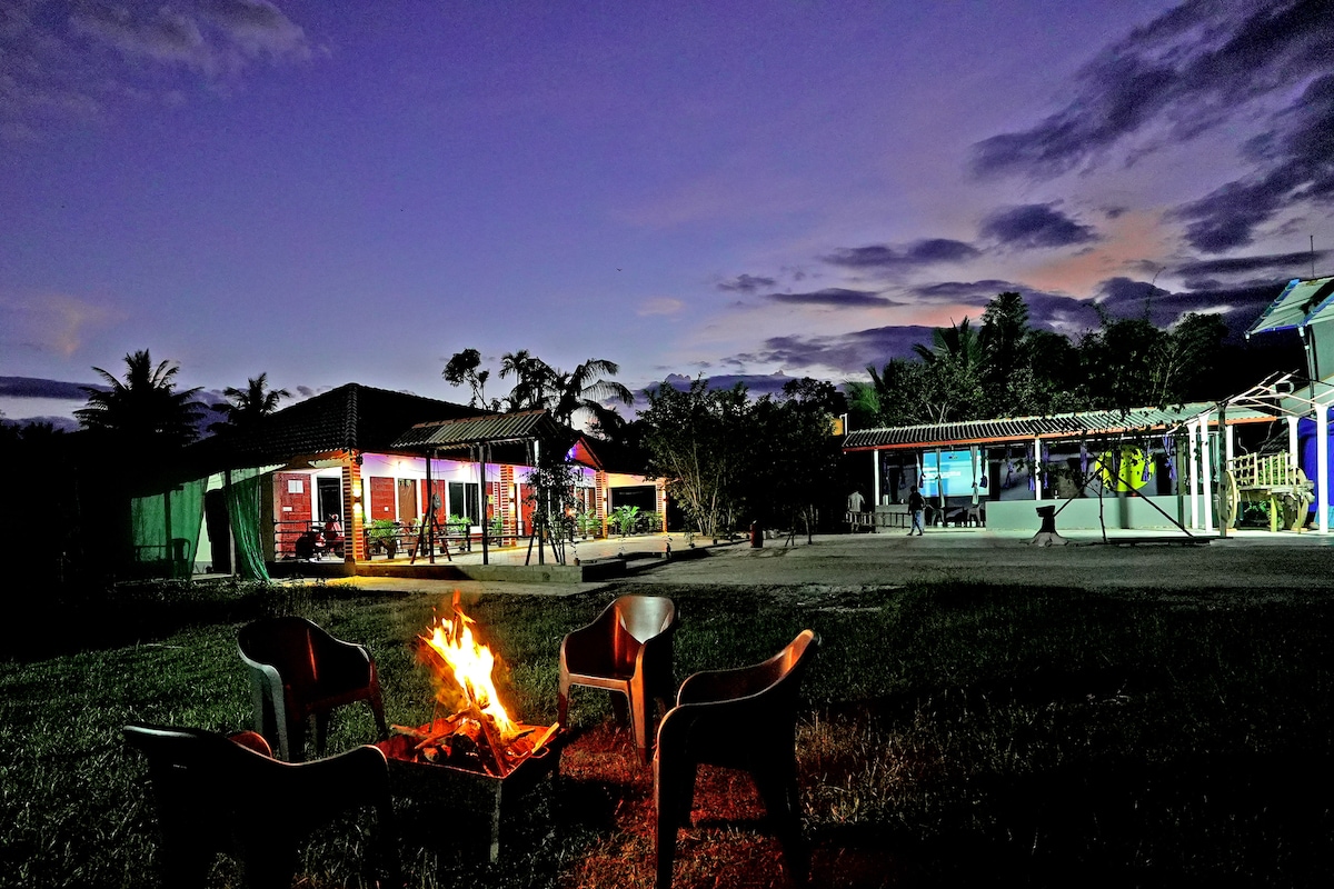 A campfire is surrounded by several chairs, providing a cozy outdoor gathering space. The background features cosy cottages illuminated by soft lighting, amidst a serene landscape under a twilight sky with scattered clouds.