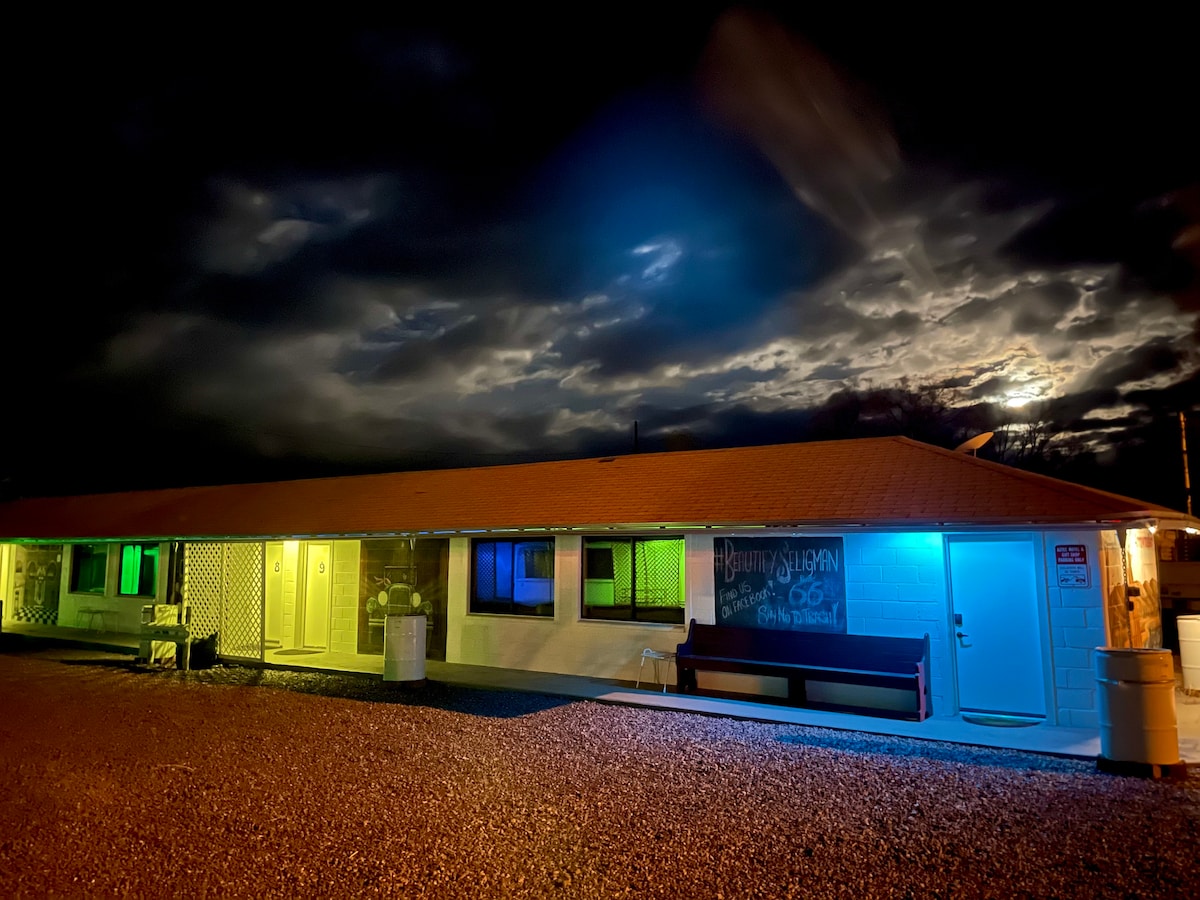 A single-story motel is illuminated with colorful lights in green, blue, and yellow against a dramatic night sky. Outdoor seating is visible, and partially lit windows create a warm ambiance. The textured gravel ground complements the overall scene.