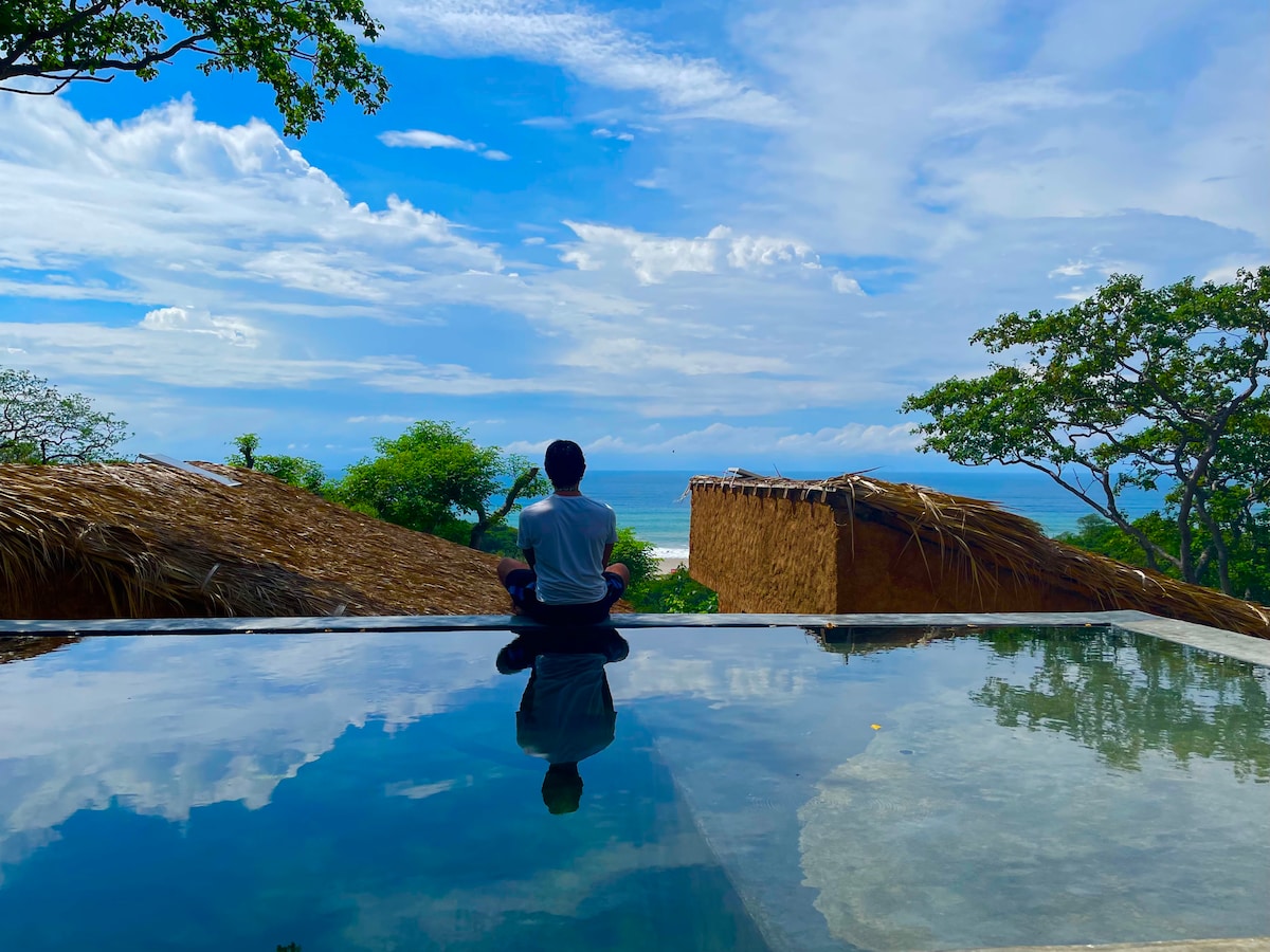 A view of a serene pool with still water reflecting the sky. In the foreground, a person sits in a meditative pose, gazing at the ocean and distant hills under a partly cloudy sky. Tropical greenery surrounds the area.