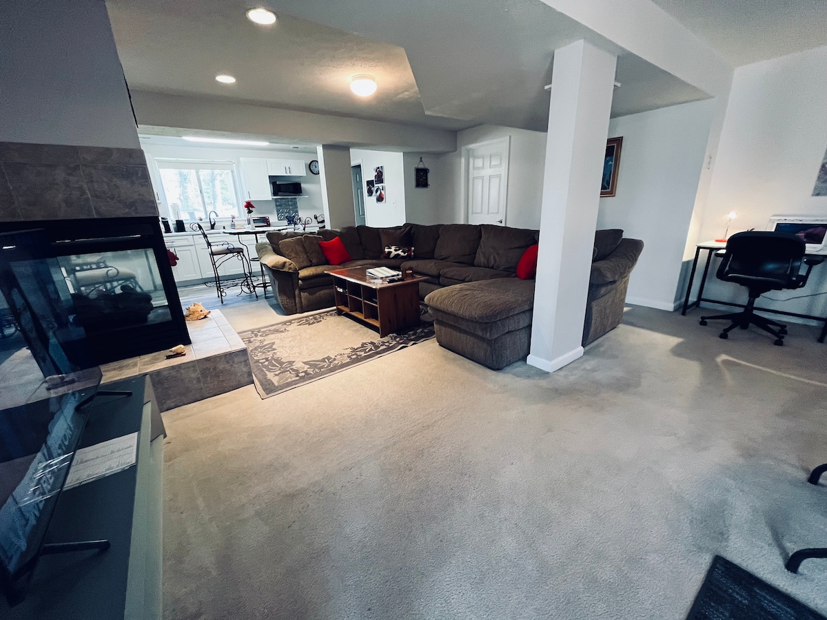A spacious living area features a large sectional sofa adorned with red accent pillows. A simple coffee table is positioned centrally, with a small work-space visible in the background. A radiant fireplace stands to the left, complementing the light-colored walls and carpet.