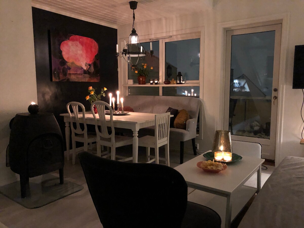 A cozy living area is illuminated by soft candlelight, featuring a dining table surrounded by white chairs and a sofa adorned with cushions. A large artwork is displayed on the wall, while a door opens to a small outdoor space. Warmth is added by a fireplace in the corner.