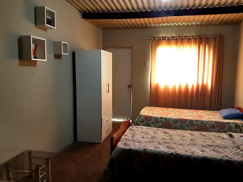 The room features two single beds dressed in floral quilts, with a small nightstand beside each. A wardrobe stands against one wall, and wall-mounted shelves display framed photos. Natural light filters through curtains, creating a bright and airy atmosphere.