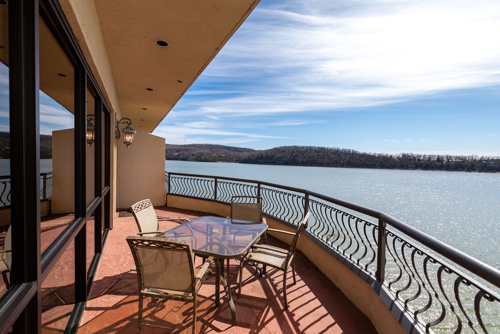 A spacious balcony features comfortable outdoor seating around a glass-top table, offering panoramic views of the peaceful lake and surrounding landscape. The gentle curves of the railing enhance the open feel, while large windows reflect natural light from the sky above.