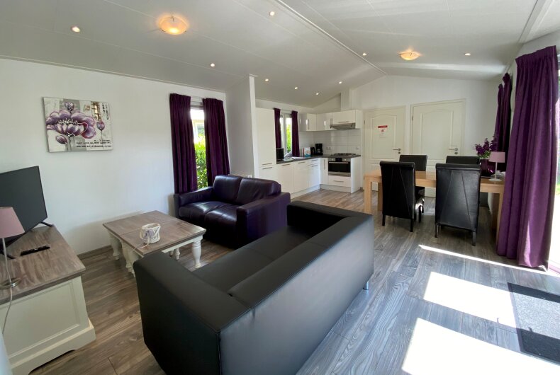 An open living area features a comfortable dark sofa and a light-colored coffee table. A dining setup with dark chairs is positioned next to a well-equipped kitchen. Large windows invite natural light, while curtains add a touch of color to the space.