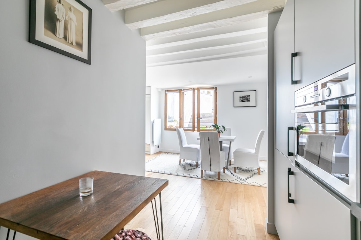 A view from the kitchen area reveals an open space leading to a dining area with white chairs around a round table. Natural light floods in through multiple large windows, illuminating the warm wooden flooring and subtle wall art.