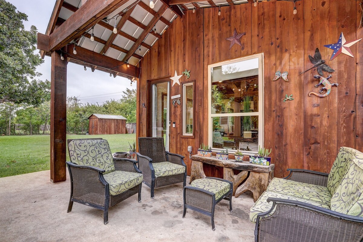 An outdoor patio area features comfortable seating with patterned cushions. Natural wood accents and decorative items adorn the wall. Lush greenery is visible in the background, adding a serene touch to the space.