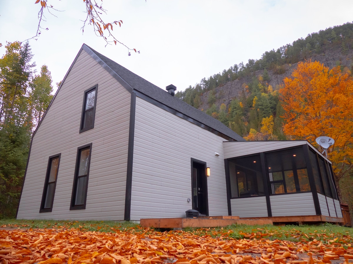 The modern chalet features a distinctive A-frame structure with a light exterior and large windows. Surrounding autumn foliage presents vibrant orange and yellow leaves. A screened porch extends from the building, providing additional outdoor living space, with a wooden deck partially visible.