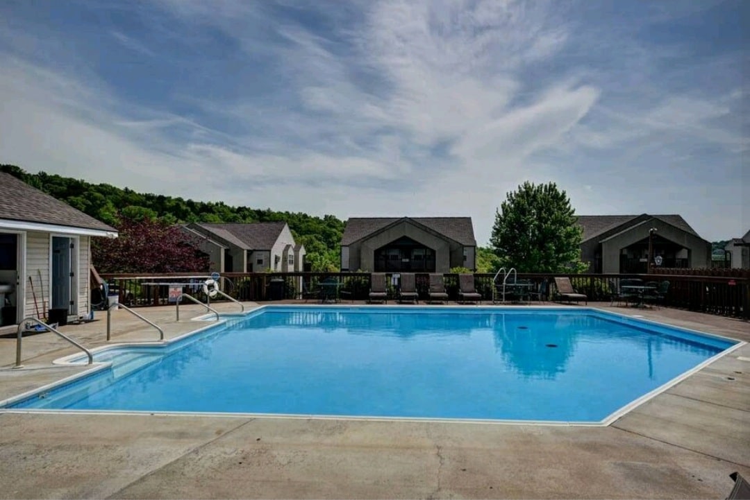 An outdoor swimming pool is depicted, featuring a clear blue surface reflecting the sky. Surrounding the pool, lounge chairs are arranged on a sun deck. A few trees and buildings can be seen in the background, adding to the serene environment.