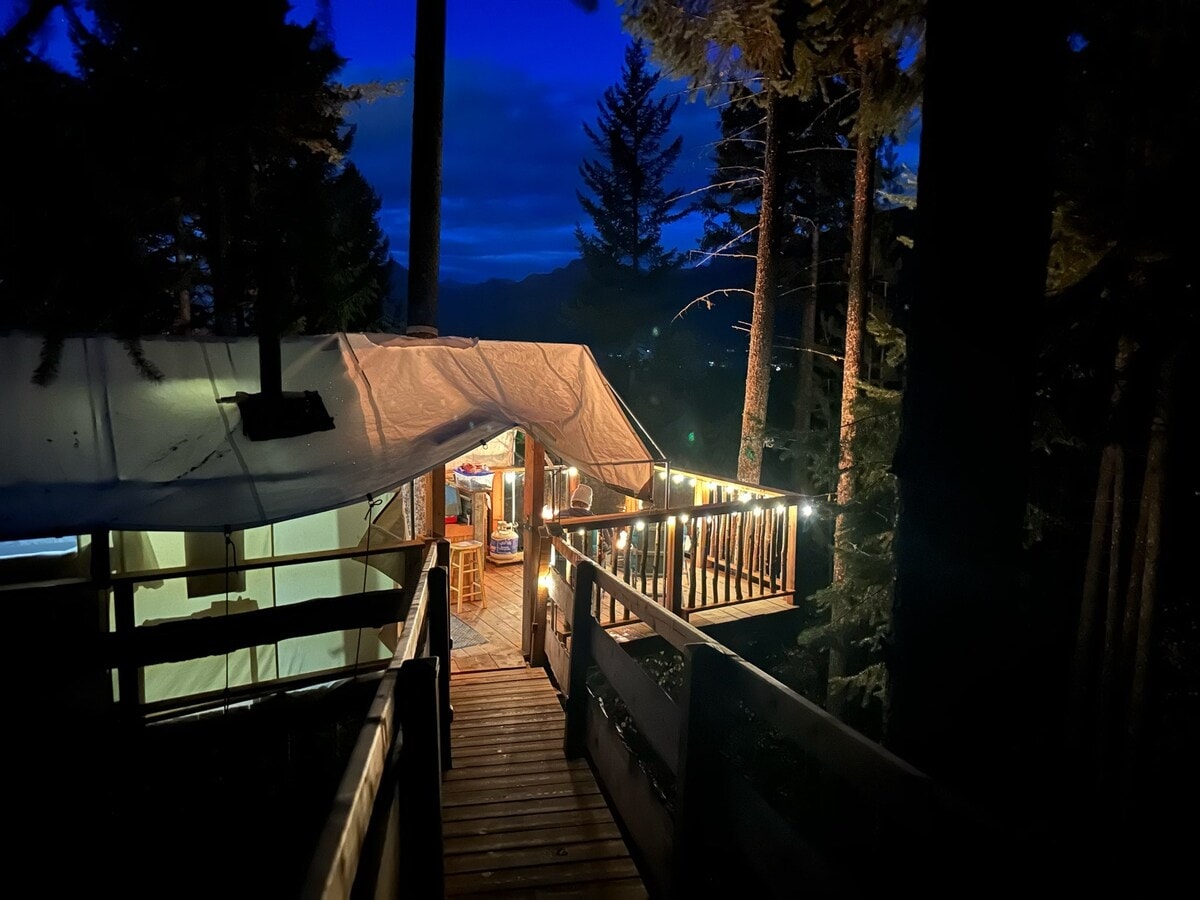 The entrance to the treehouse tent is illuminated by string lights, casting a warm glow. Wooden decking leads into the structure, surrounded by tall trees under a twilight sky. Soft blue hues create a serene atmosphere as night begins to settle in.