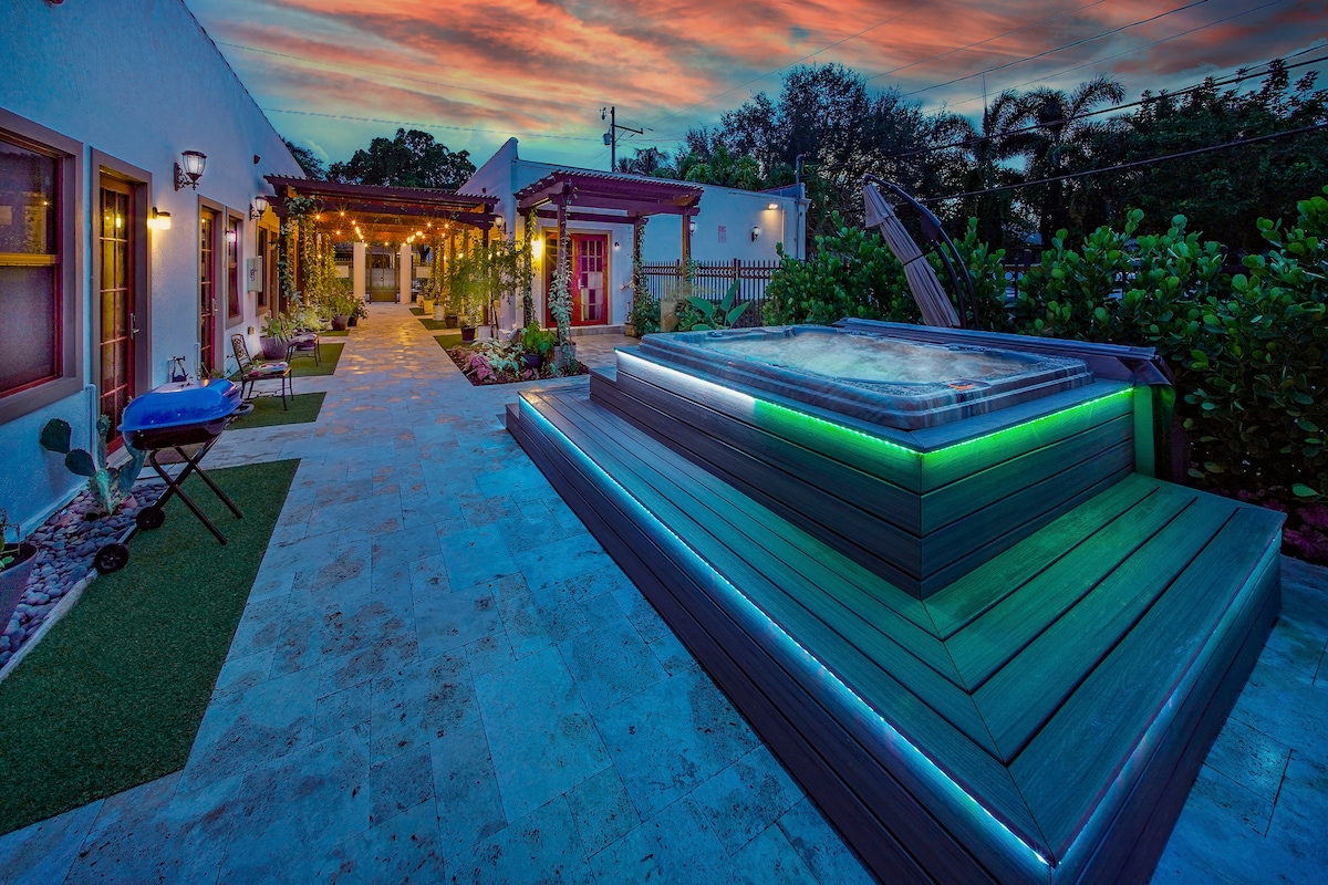 An inviting outdoor area showcases a well-lit hot tub surrounded by elegant landscaping. Warm sunset hues paint the sky, while soft lighting illuminates the pathways and foliage, creating a serene atmosphere for relaxation and leisure.